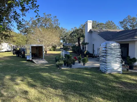 Shrubs and palms in pots and stacked bags of mulch on a patio, with the work trailer parked on the lawn